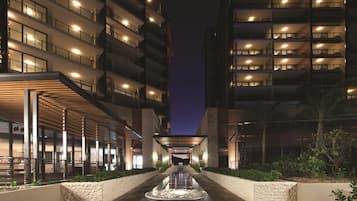 A nighttime view of a hotel entrance with a central water feature, multiple floors with illuminated windows, and a covered walkway on either side.