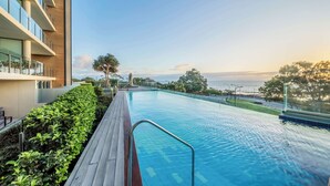 A modern hotel pool with a wooden deck, surrounded by lush greenery and offering a view of the sea.