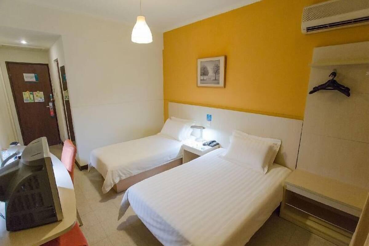 A hotel room with two beds, a desk with a telephone, a hanging light fixture, a wall-mounted air conditioner, and a framed picture.