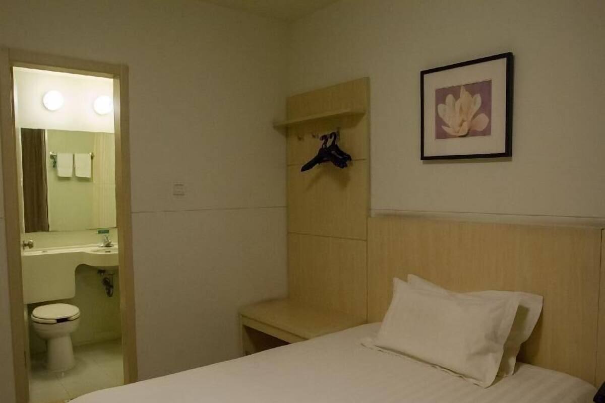A hotel room with a bed, a bedside table, a wall-mounted shelf with a coat hanger, a framed picture, and a bathroom visible through an open door.