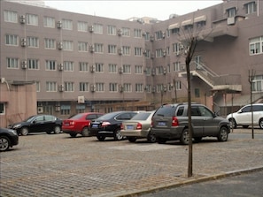 A parking area with several cars parked in front of a multi-story building with multiple windows and air conditioning units.