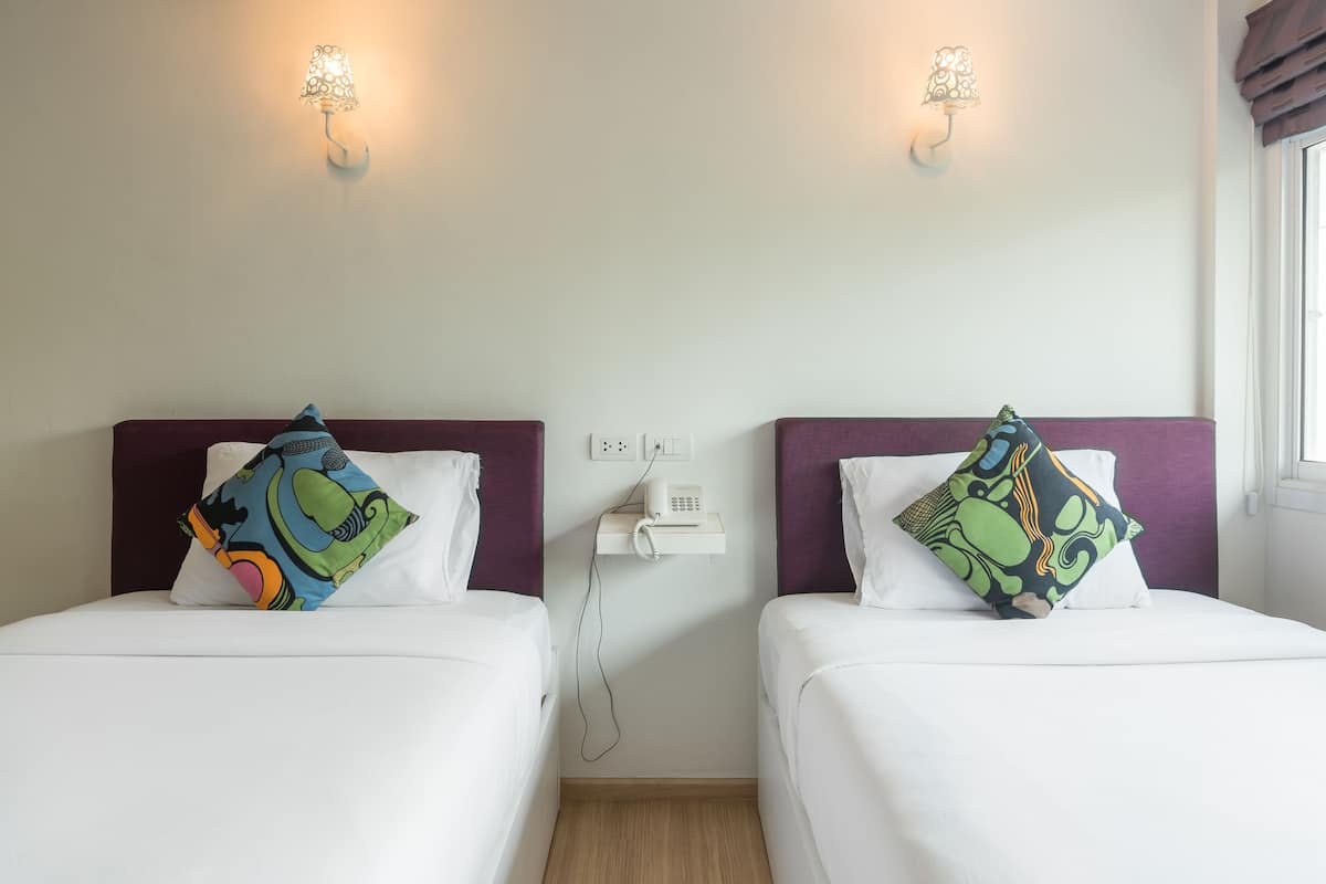 Deux lits simples avec du linge de lit blanc, des têtes de lit violettes et des oreillers colorés. Des lampes murales et un téléphone sont posés sur une étagère.