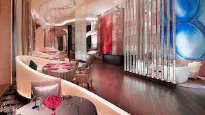 En luksuriøs hotellobby med et moderne design, der inkluderer et centralt siddeområde med runde borde og stole, et stort abstrakt vægmaleri og en dekorativ søjle med hængende detaljer.