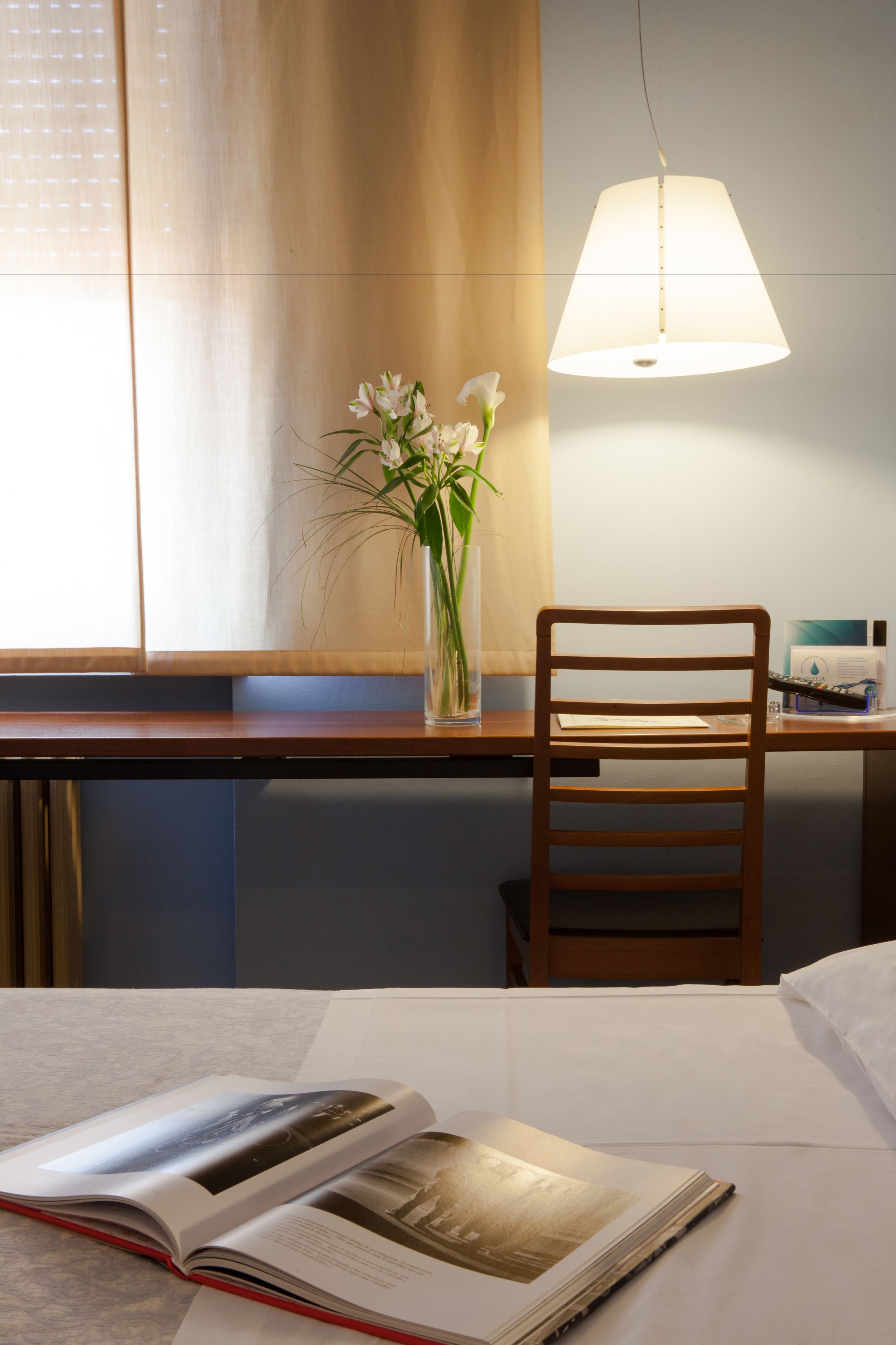Une chambre d’hôtel avec un bureau en bois, une chaise, une table de chevet avec un livre et une lampe.