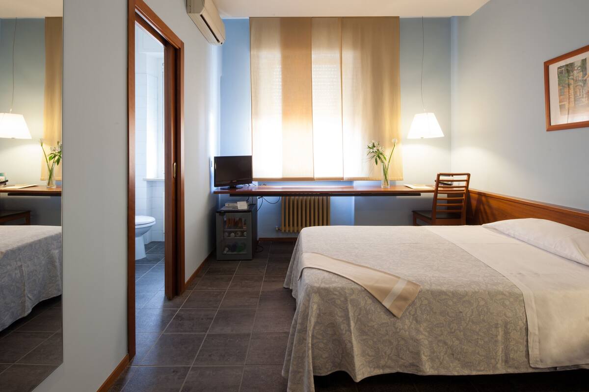 Habitación de hotel con dos camas, un escritorio con computadora, una silla y un ventanal con cortinas.