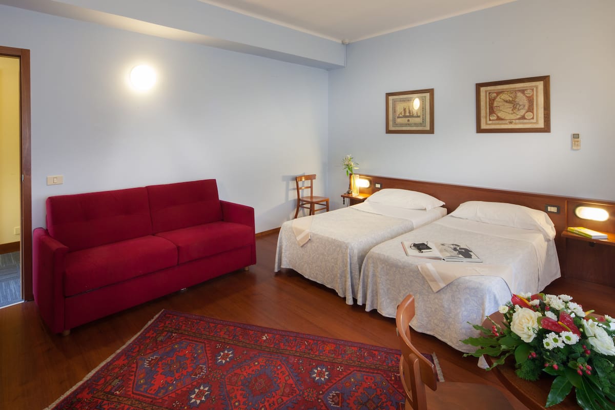 Habitación de hotel con dos camas, un sofá rojo, una silla de madera, una mesita de noche con lámpara y un arreglo floral.