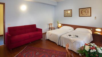 Habitación de hotel con dos camas, un sofá rojo, una silla de madera, una mesita de noche con lámpara y un arreglo floral.