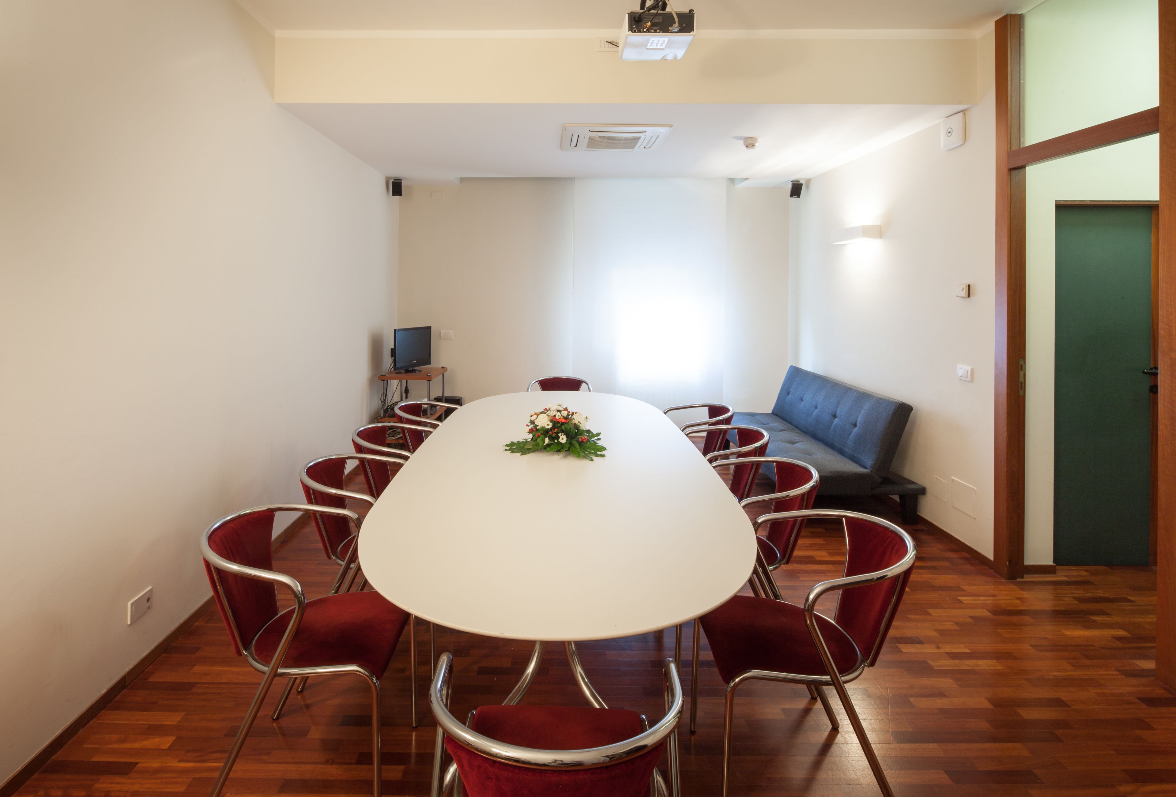 A conference room with a white table, red chairs, a blue sofa, and a television on a stand.