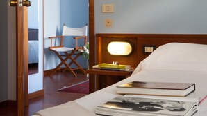 A hotel room with a bed, a bedside table with books, a chair, and a nightstand with a lamp.