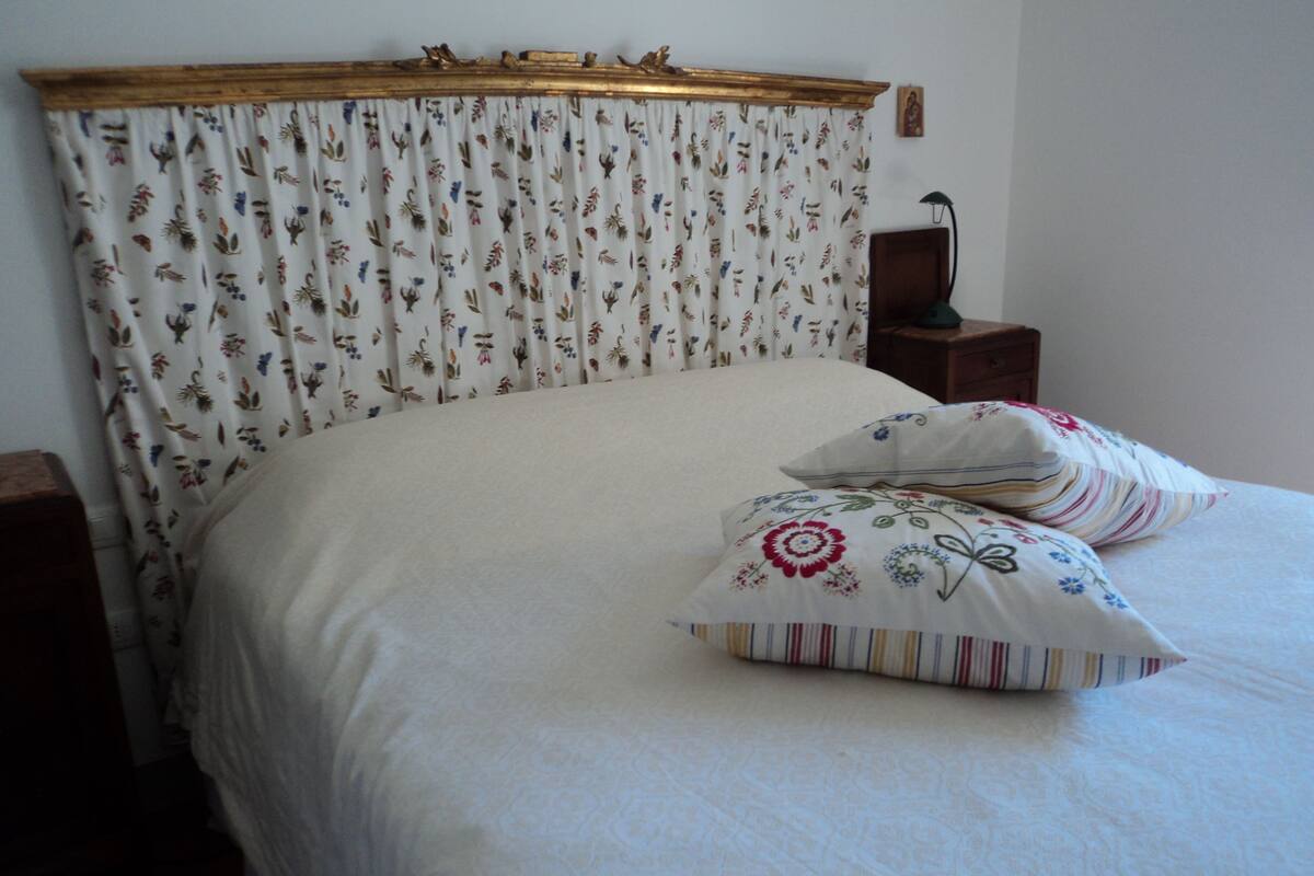 A neatly made bed with floral-patterned bedding and pillows, a wooden headboard, and a small bedside table with a lamp.