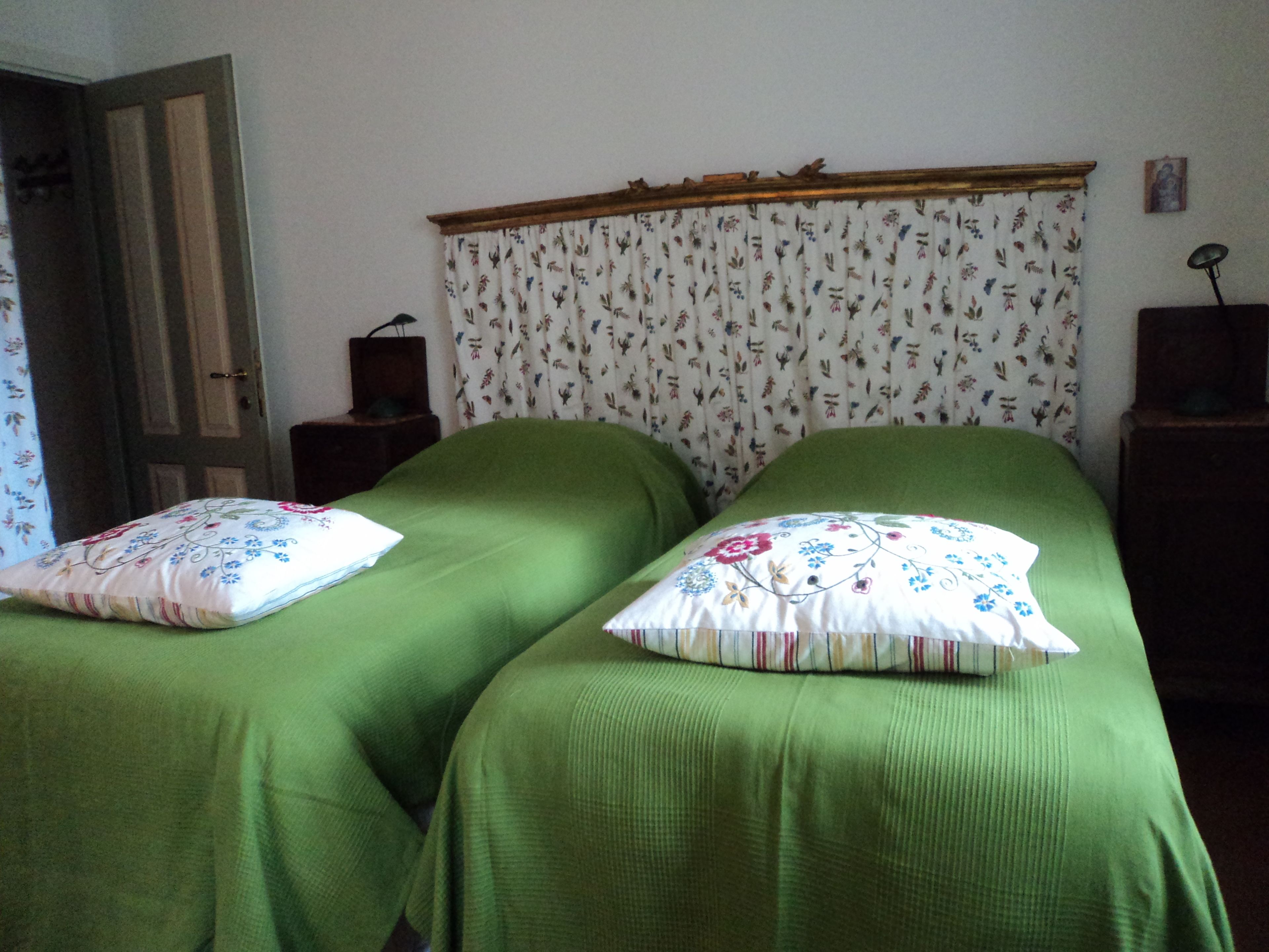 Habitación con dos camas individuales, cada una con cubrecama verde y almohada estampada. Se observa un cabecero de madera, una mesita con lámpara y una puerta con cortina floral.