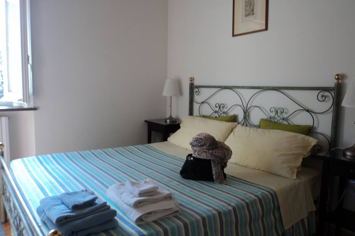 A neatly made bed with striped bedding, a lamp on the nightstand, and folded towels on the floor.