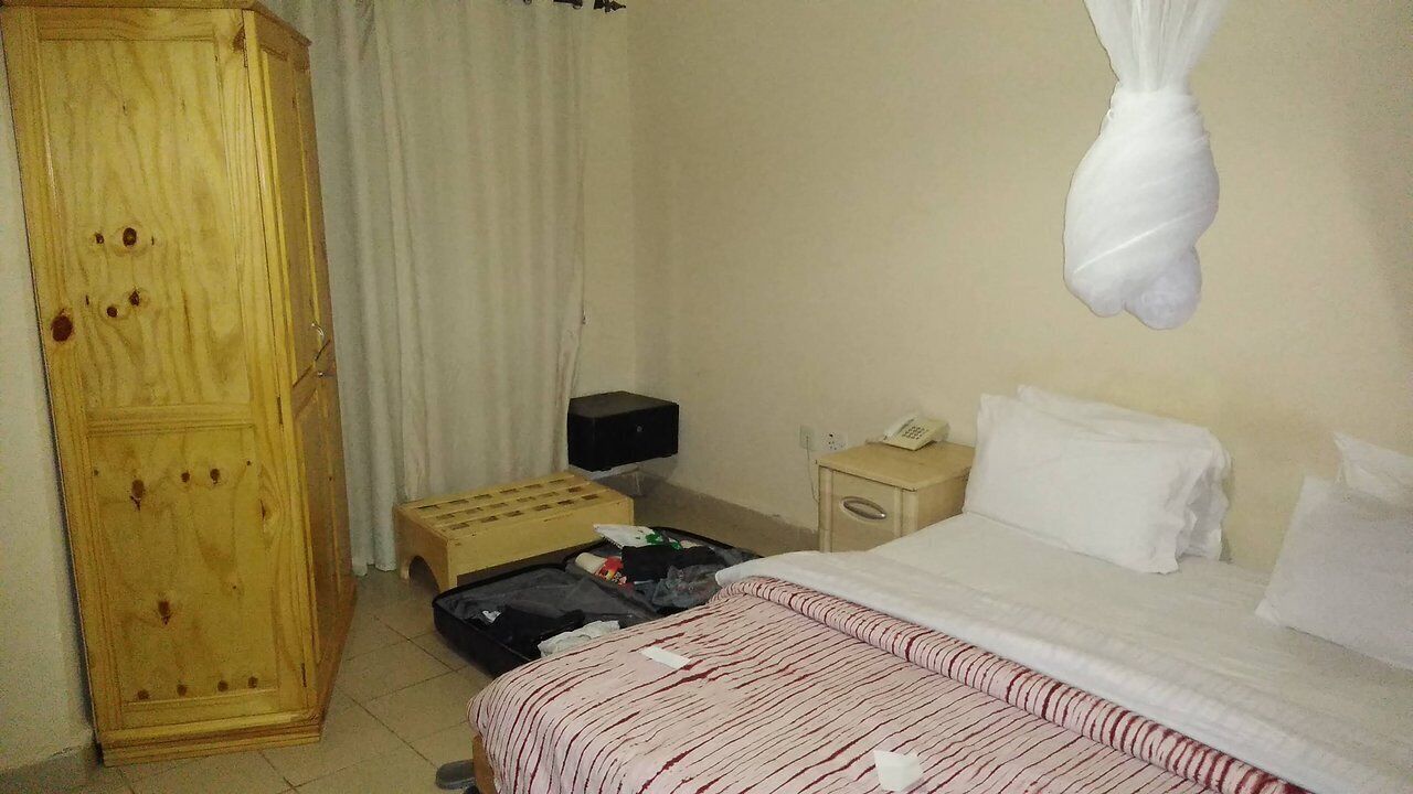 A hotel room with a wooden wardrobe, a bed with striped bedding, a bedside table with a telephone, and a hanging white object.