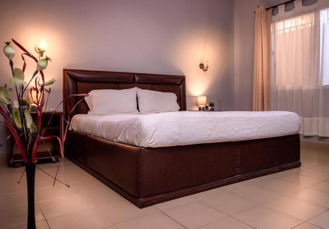 Una cama bien tendida con cabecera de madera, una mesita de noche con lámpara y una planta en maceta a la izquierda.