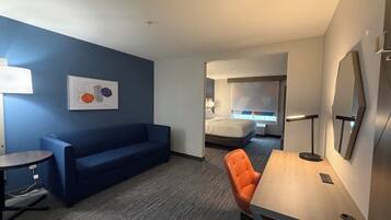 A hotel room with a blue sofa, an orange chair, a wooden table, a bed with a white headboard, and a large mirror.