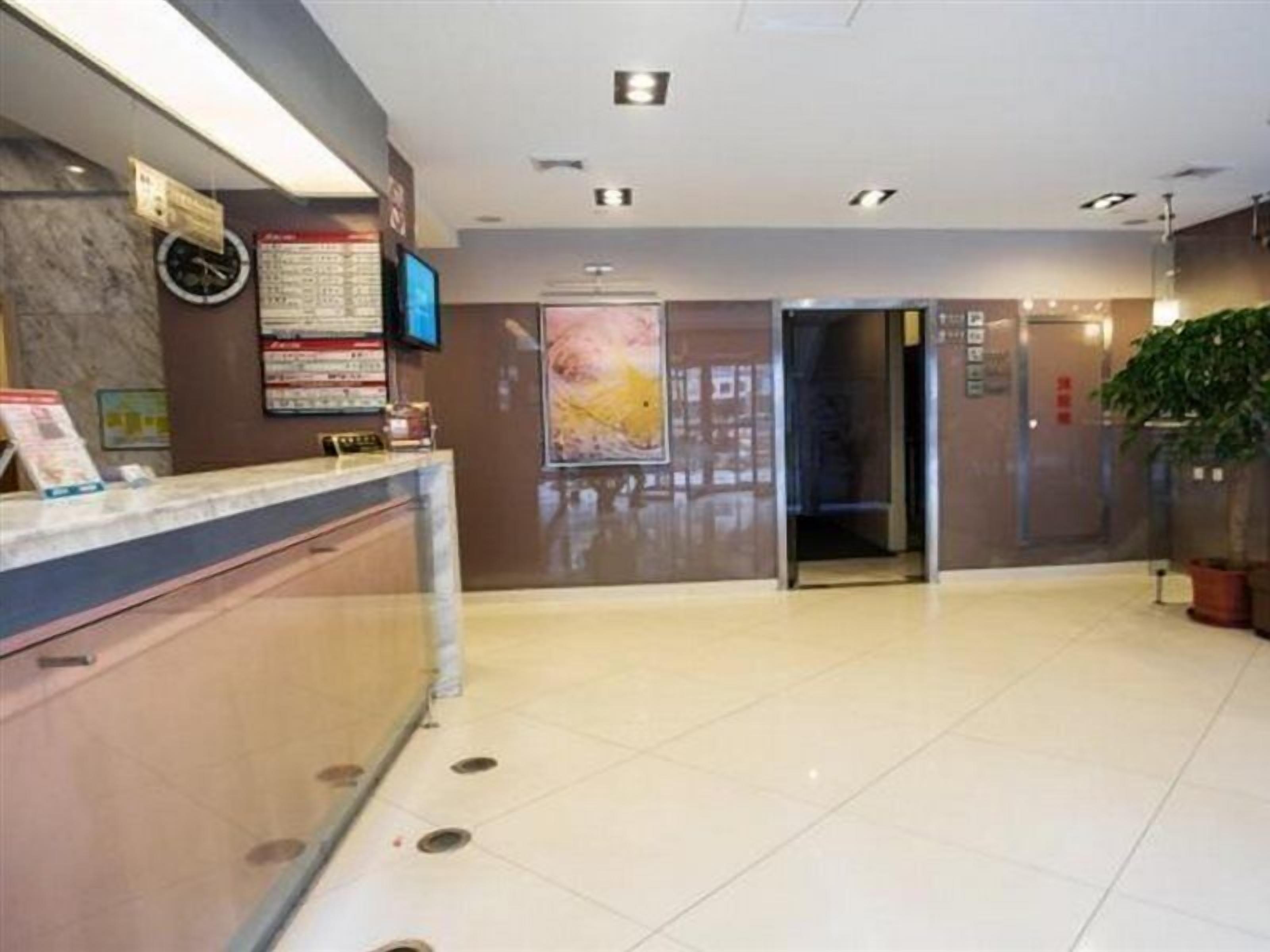 Zona de recepción de un hotel con mostrador, un reloj, una pantalla digital, un cartel y un ascensor.