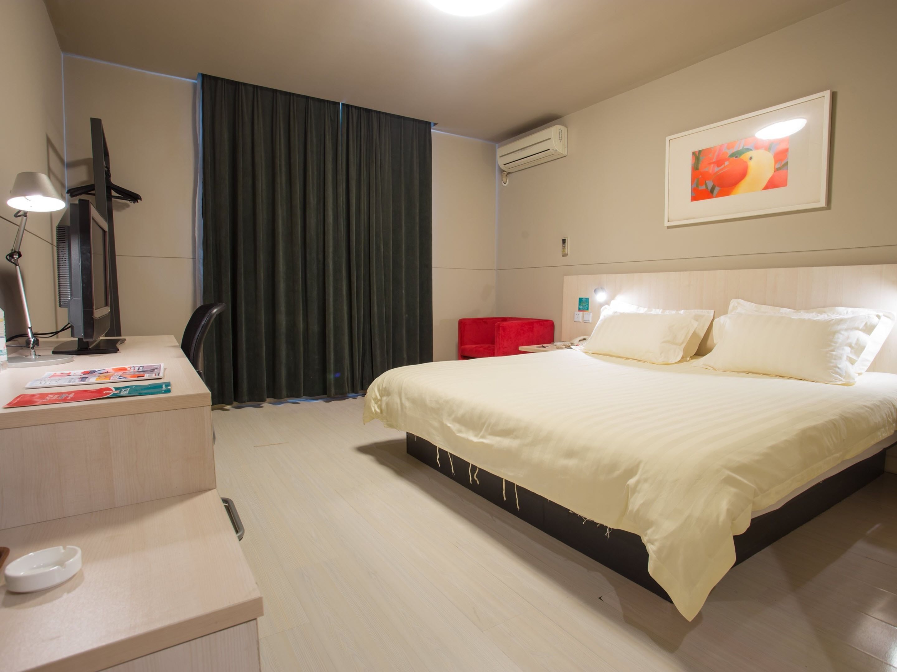 A hotel room with a bed, a desk with a lamp, a chair, a red chair, a painting on the wall, and an air conditioning unit.