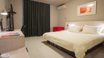 A hotel room with a bed, a desk with a lamp, a chair, a red chair, a painting on the wall, and an air conditioning unit.