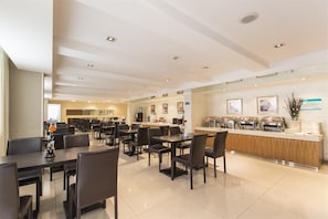 Een moderne eetruimte van een hotel met meerdere tafels en stoelen, een balie met horeca-apparatuur, en lijstwerken aan de muren.