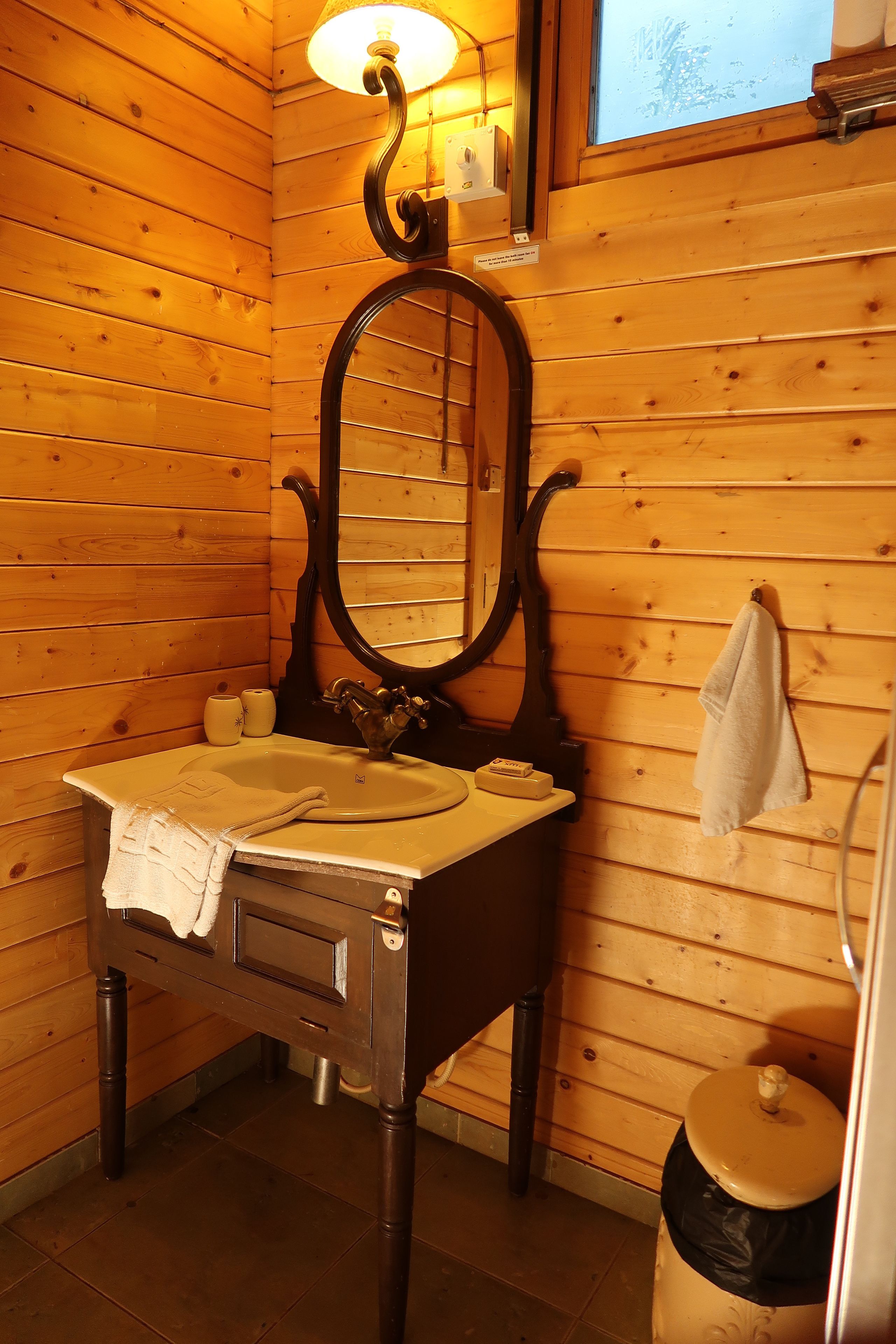 A wooden cabin bathroom with a wooden sink, a mirror, a towel hanging on the wall, and a window above the mirror.