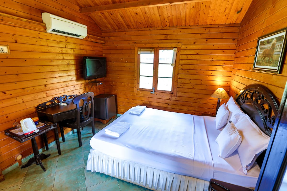 A wooden cabin room with a bed, a desk, a chair, a television, and a window.