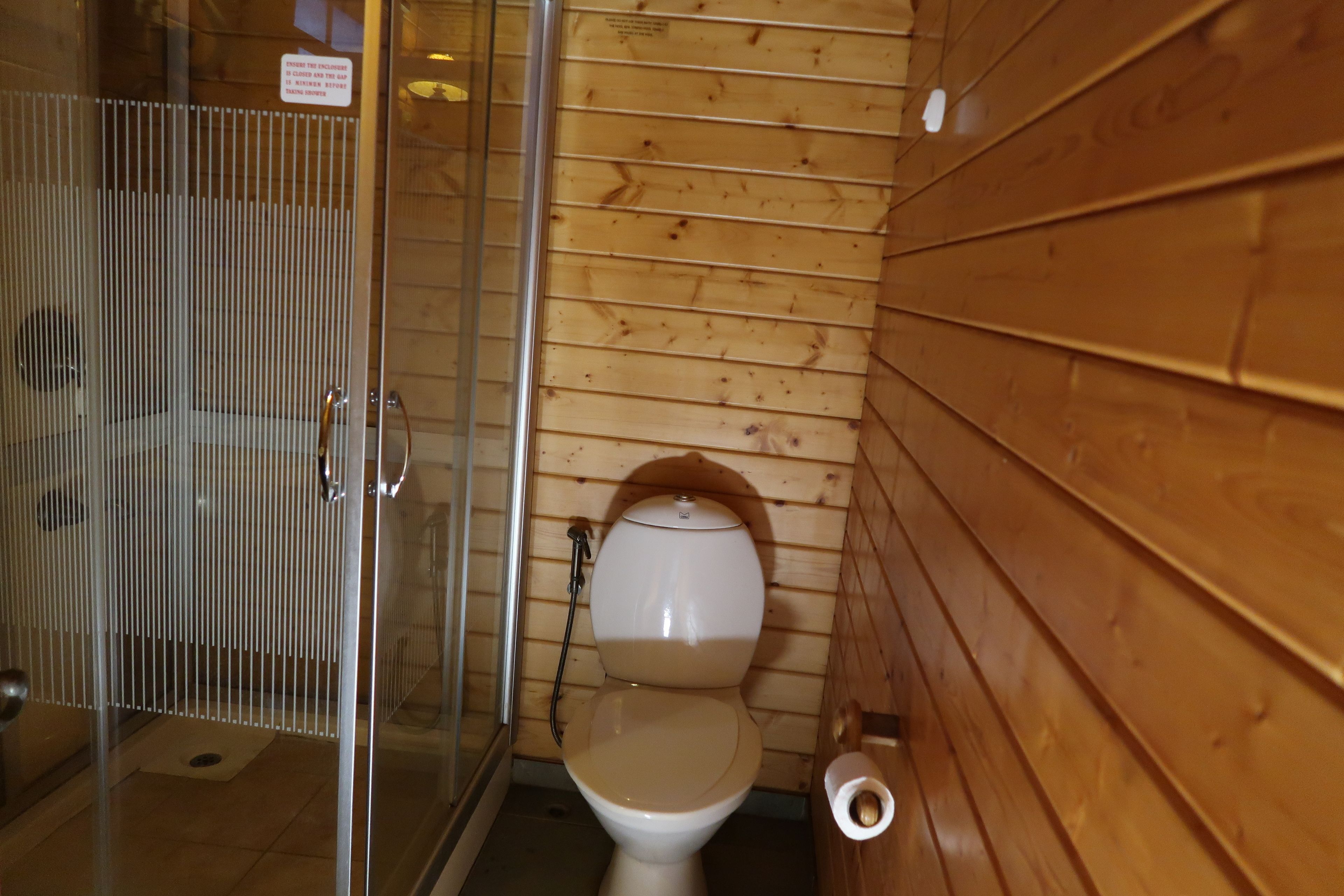 A small, compact bathroom with a wooden wall, a toilet, a shower enclosure, and a toilet paper holder.