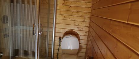 A small, compact bathroom with a wooden wall, a toilet, a shower enclosure, and a toilet paper holder.