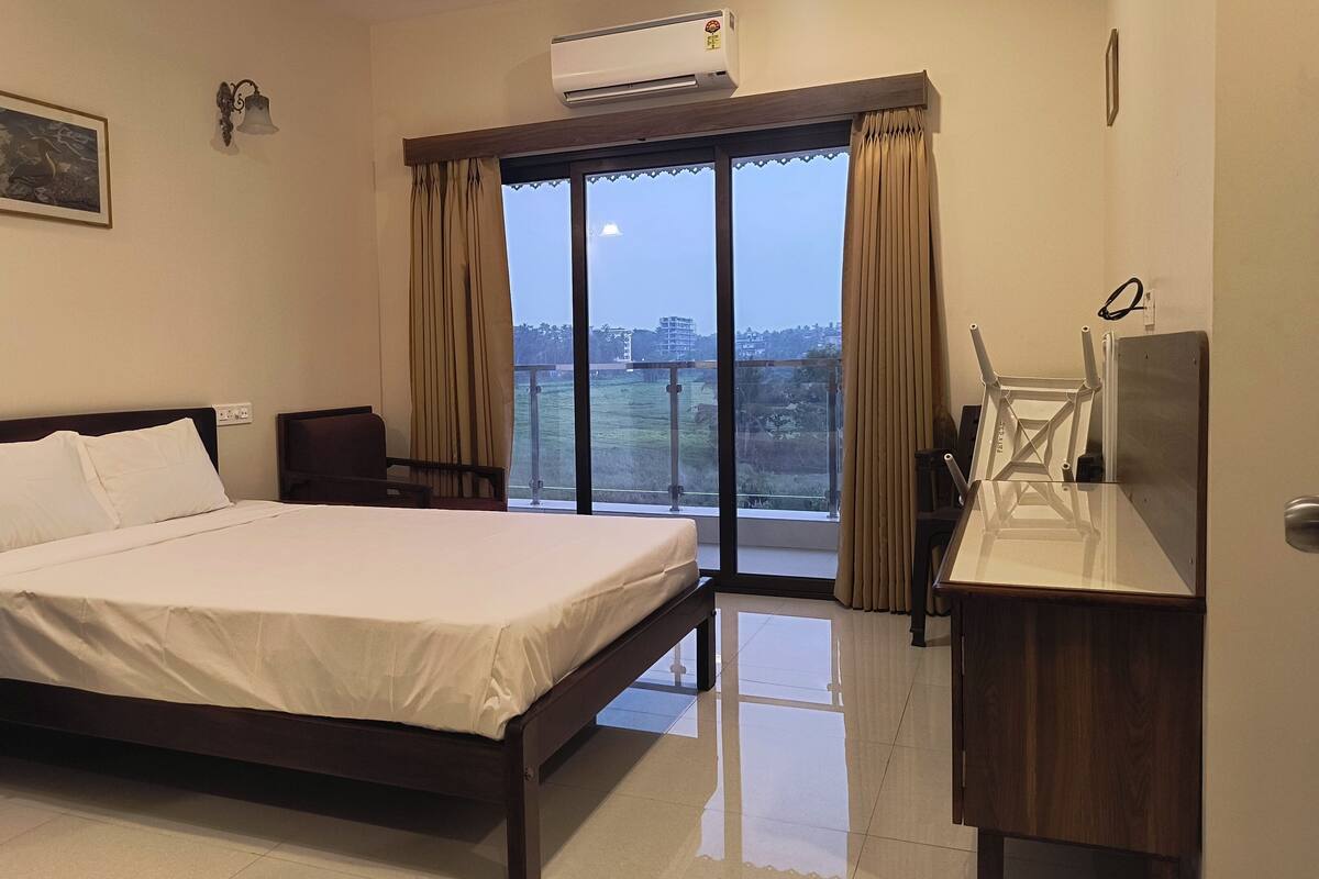A hotel room with a large bed, a desk with a mirror, an air conditioner, and a view of the city through the window.
