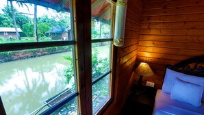 A wooden cabin room with a bed, a nightstand, a lamp, and a window overlooking a water body. - Resort Primo Bom Terra Verde (Baga)