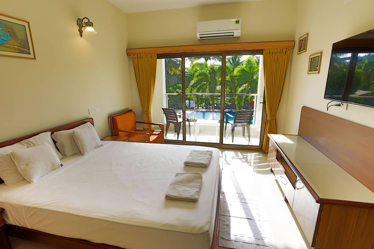 A hotel room with a large bed, a desk, a chair, a television, and a view of a swimming pool and palm trees through the window.