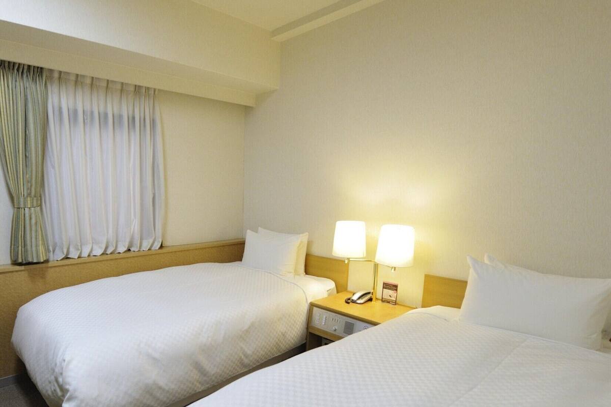 Une chambre d’hôtel avec deux lits, une table de chevet avec une lampe et une fenêtre avec des rideaux.