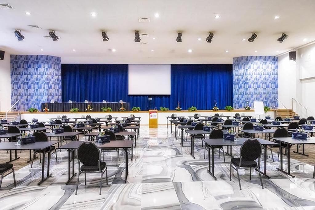 Une salle de conférence équipée d’une scène, de rangées de tables et de chaises.