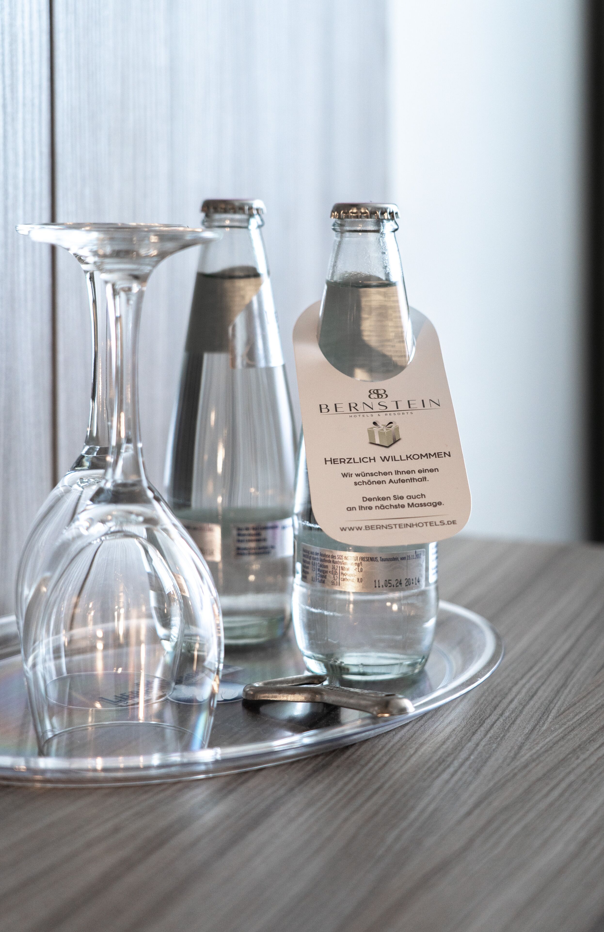 Una botella de agua del hotel Bernstein, un vaso y una bandeja de metal sobre una superficie de madera.