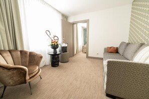 A hotel room with a brown leather armchair, a grey patterned sofa, a small table with a vase of flowers, and a view of the hallway. - Bernstein B&B (Stassfurt)