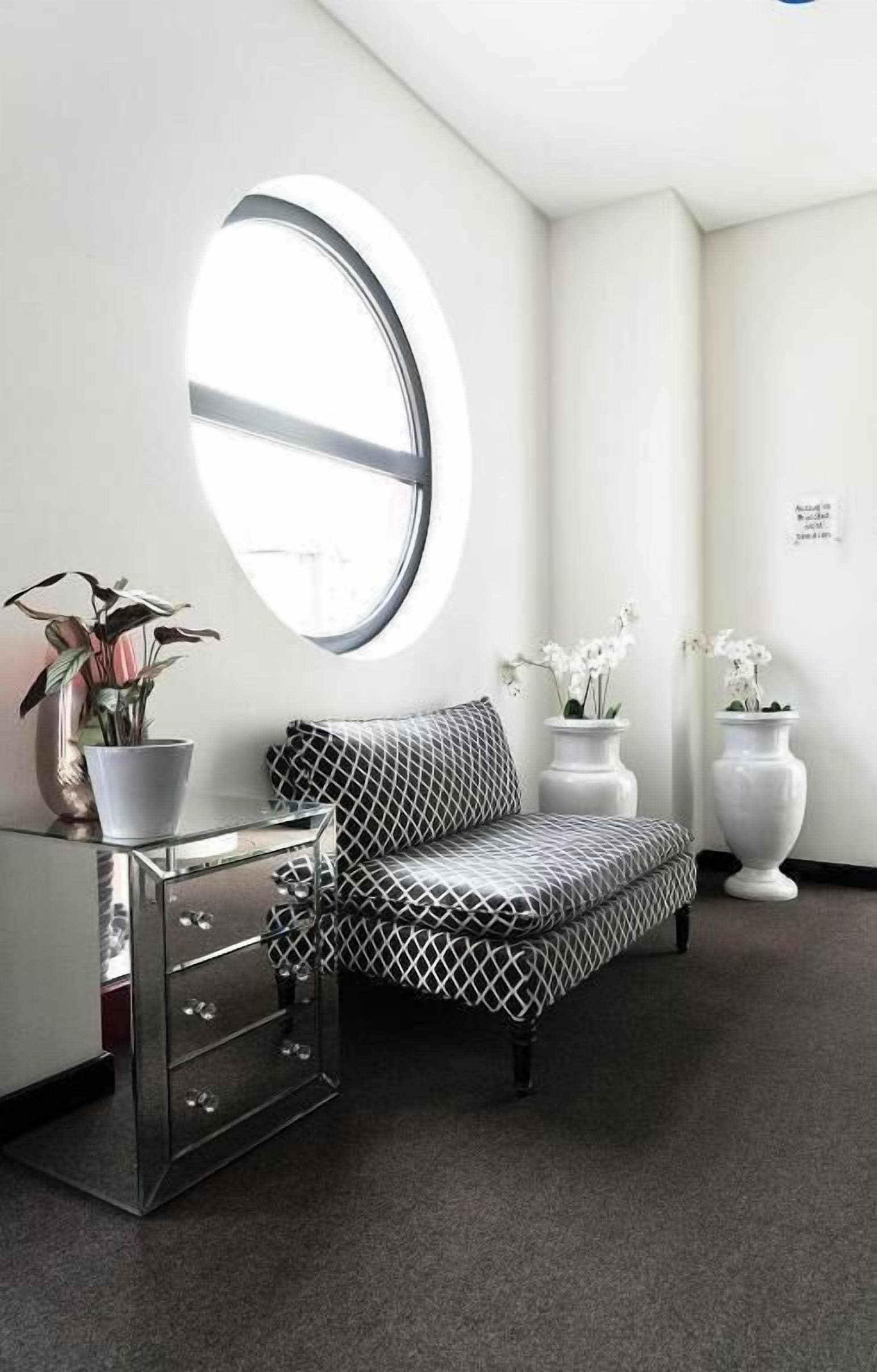 A room with a round window, a sofa, a dresser, and two potted plants.