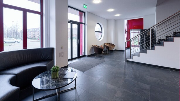 A modern lobby with a curved black sofa, a marble coffee table, a round window, and a staircase with metal railings. - Bernstein B&B (Stassfurt)