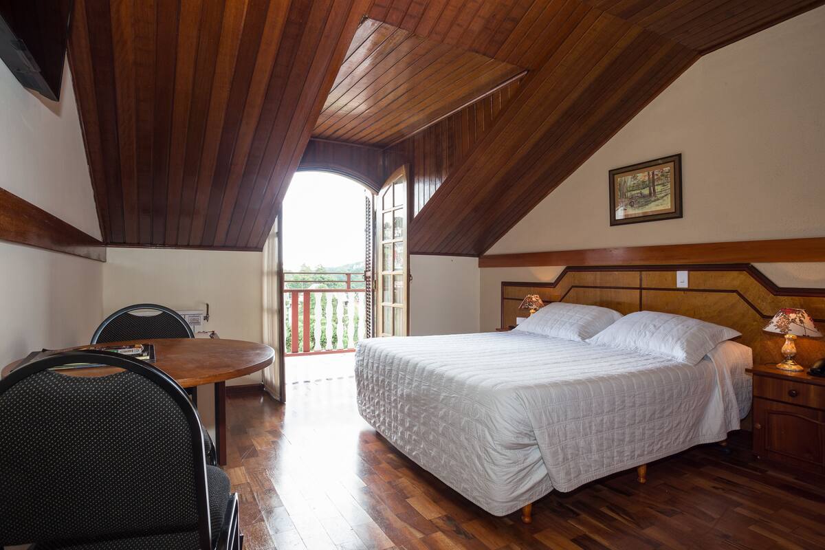 A bedroom with a wooden ceiling, a bed with white bedding, a wooden nightstand, a chair, and a table.
