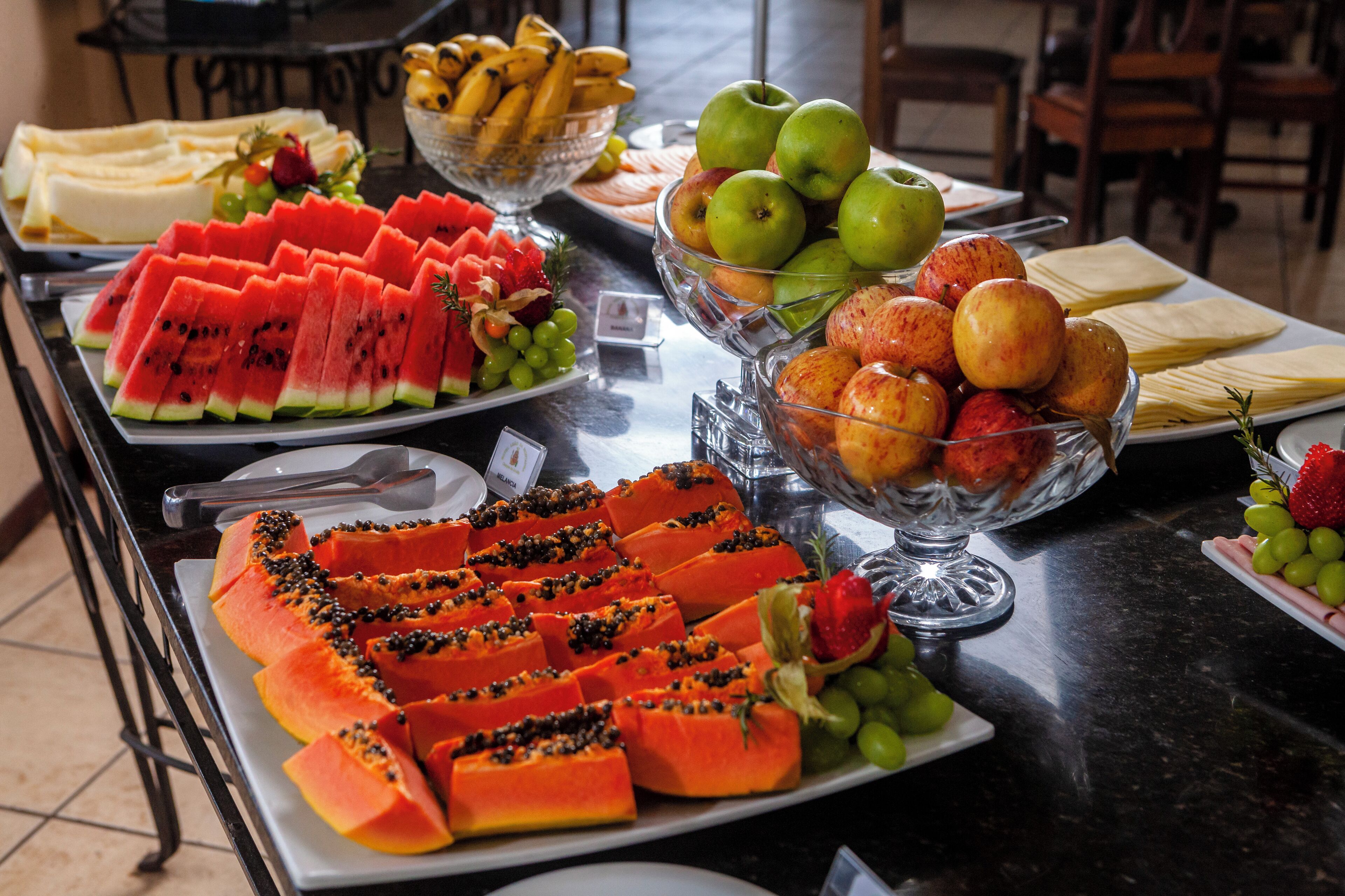 Mesa de buffet com diversas frutas e lanches, incluindo melancia, mamão, maçãs e uvas.