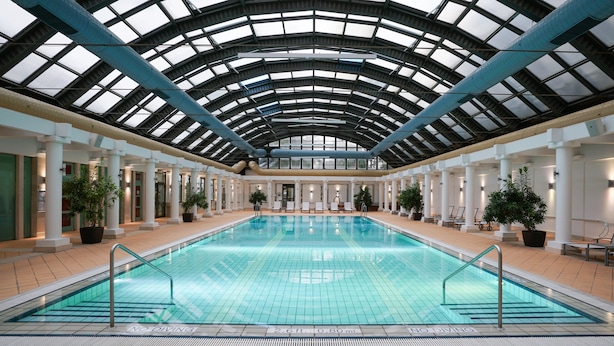 An indoor swimming pool with a glass ceiling, surrounded by columns and potted plants.
