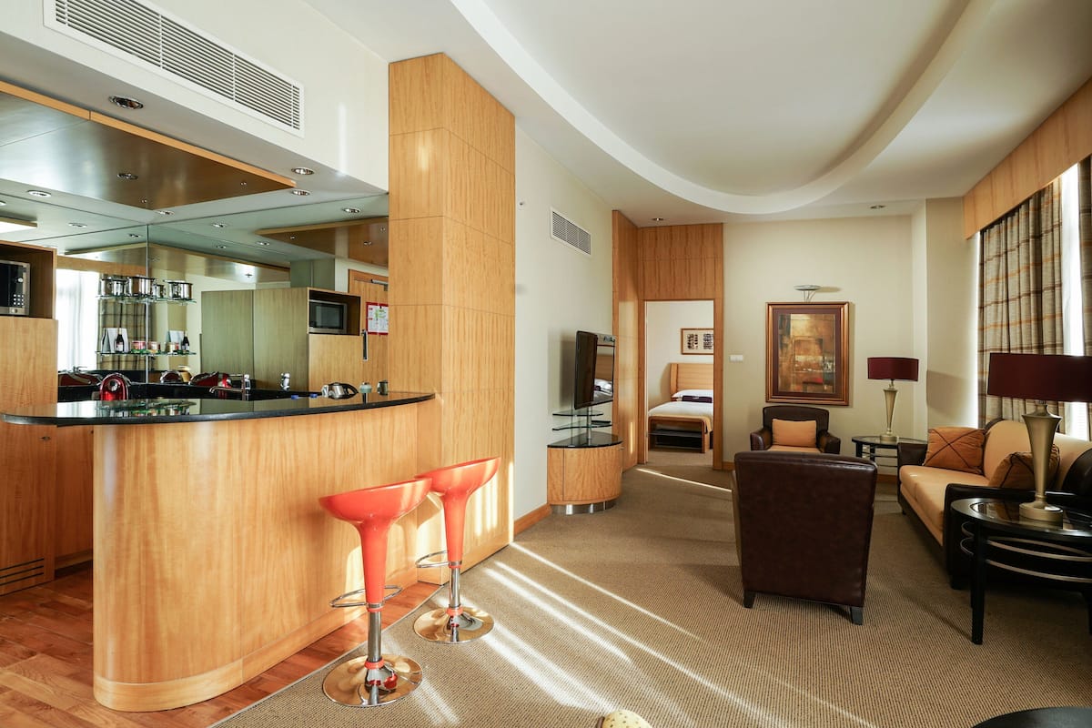 A modern hotel room with a kitchen area, a bar, and a seating area with a sofa and armchairs.