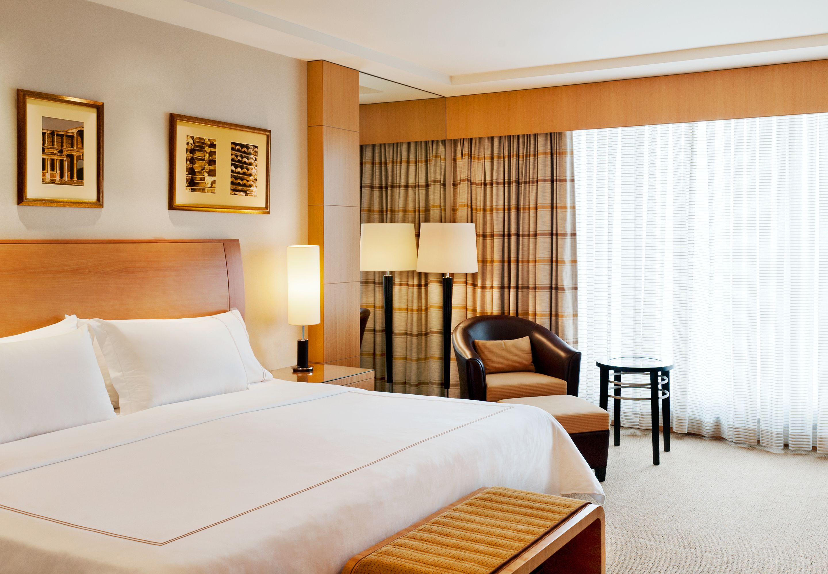 A hotel room with a large bed, two bedside lamps, a chair, a small table, and two framed pictures on the wall.