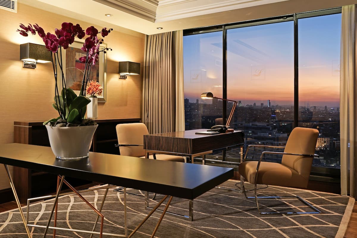 A modern hotel room with a large window offering a city view at dusk, a desk with a lamp, and a vase of orchids.