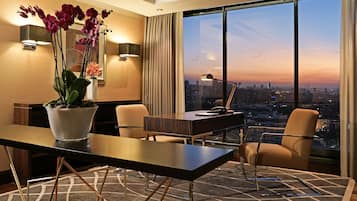 Une chambre d’hôtel moderne dotée d’une grande fenêtre offrant une vue sur la ville au crépuscule, d’un bureau avec une lampe et d’un vase rempli d’orchidées.