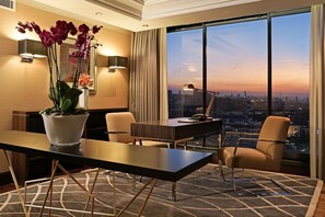 A modern hotel room with a large window offering a city view at dusk, a desk with a lamp, and a vase of orchids.