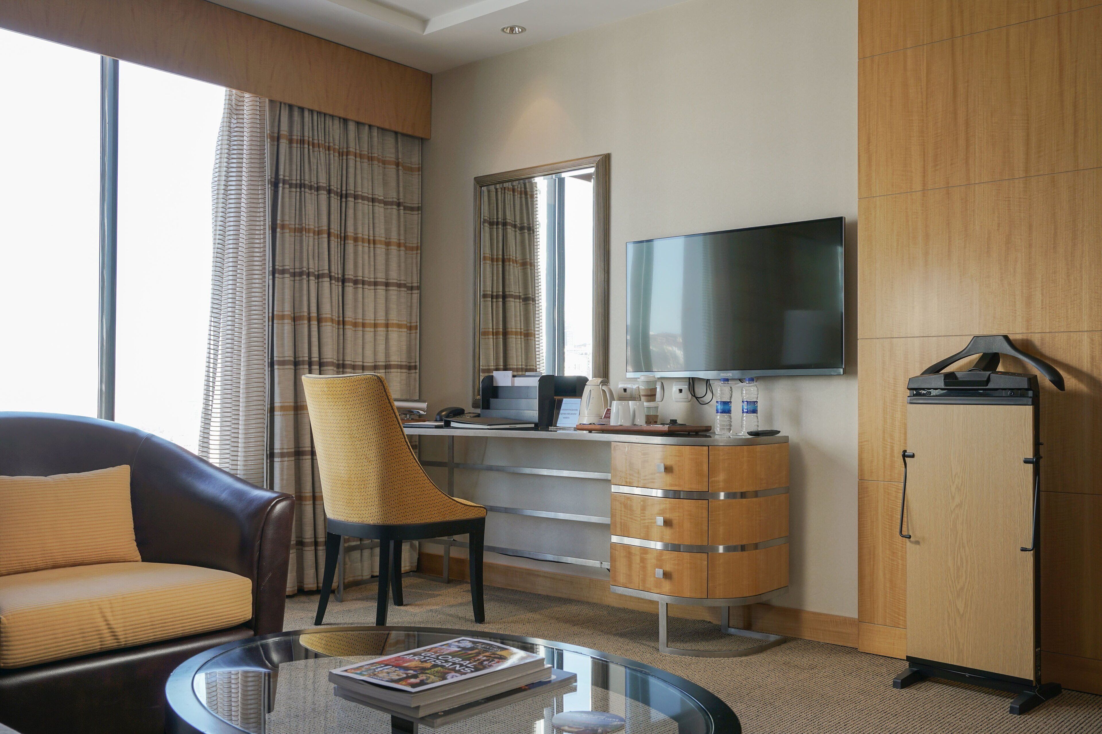 A hotel room with a sofa, armchair, coffee table, TV, wardrobe, and a view of the city through large windows.