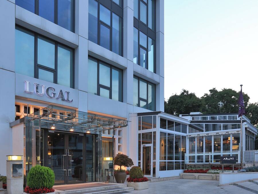 A modern hotel with a glass entrance, a sign reading "LUGAL," and a small garden area with potted plants and flowers.