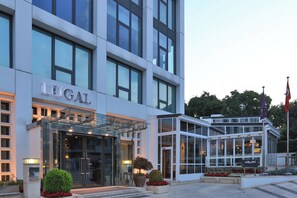 A modern hotel with a glass entrance, a sign reading "LUGAL," and a small garden area with potted plants and flowers.