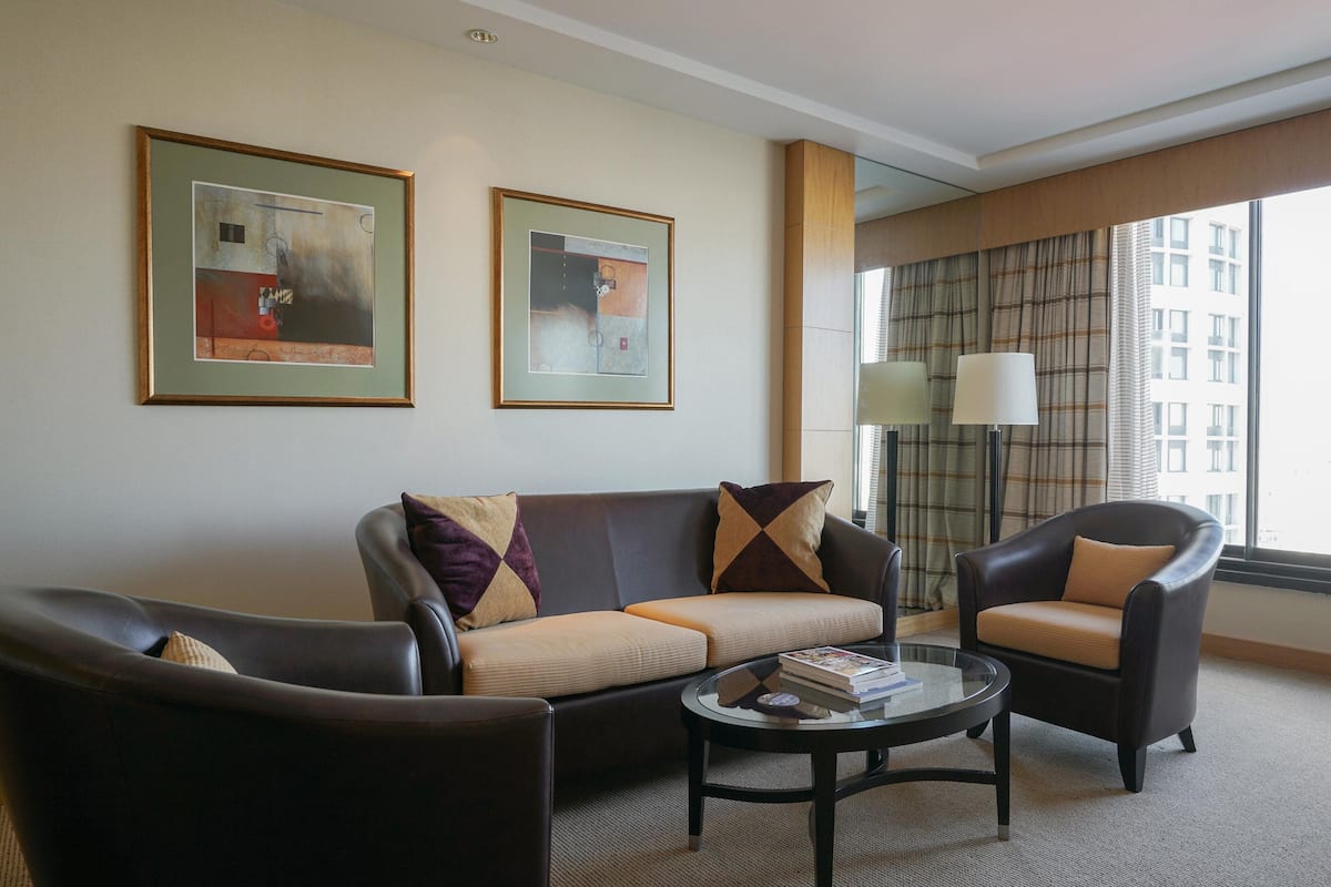 A modern hotel room with a leather sofa, armchair, and a glass coffee table. There are two framed abstract paintings on the wall and a large window with curtains.