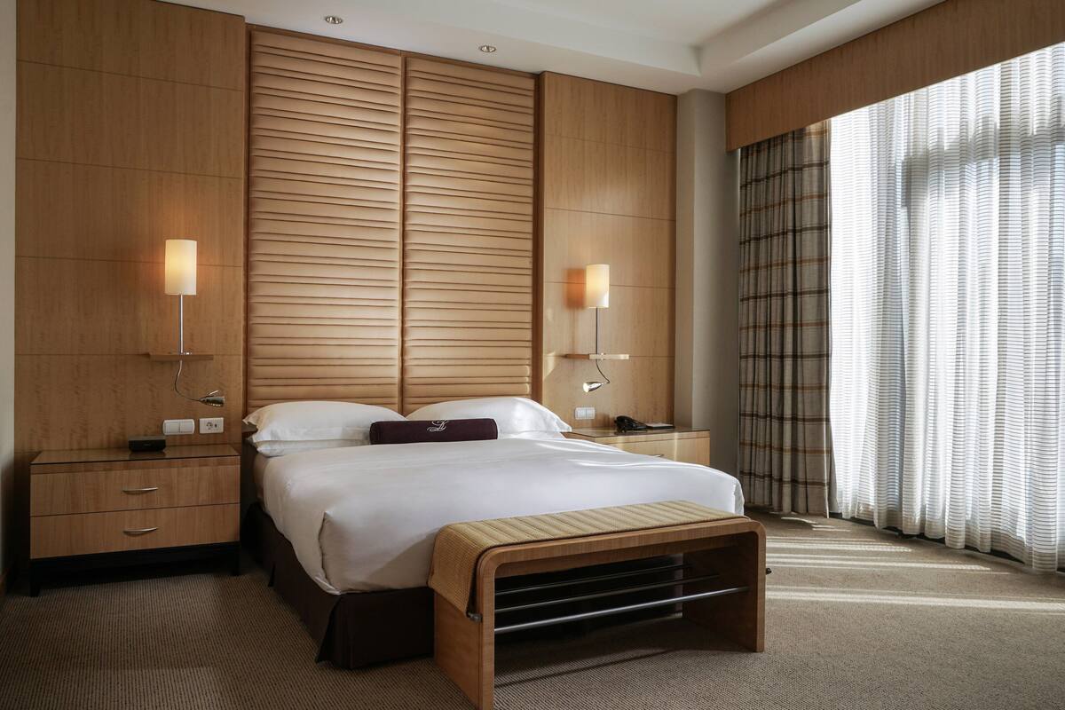 A hotel room with a large bed, wooden headboard, bedside tables with lamps, a bench, and a view of the outside through curtains.