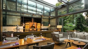 A modern outdoor dining area with wooden tables and chairs, a brick oven, and a glass-enclosed space with natural light.